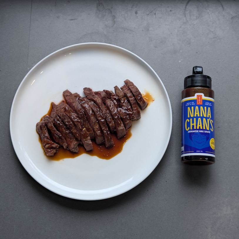Sliced steak on a white plate with a bottle of Nana-chan's BBQ sauce on a gray surface